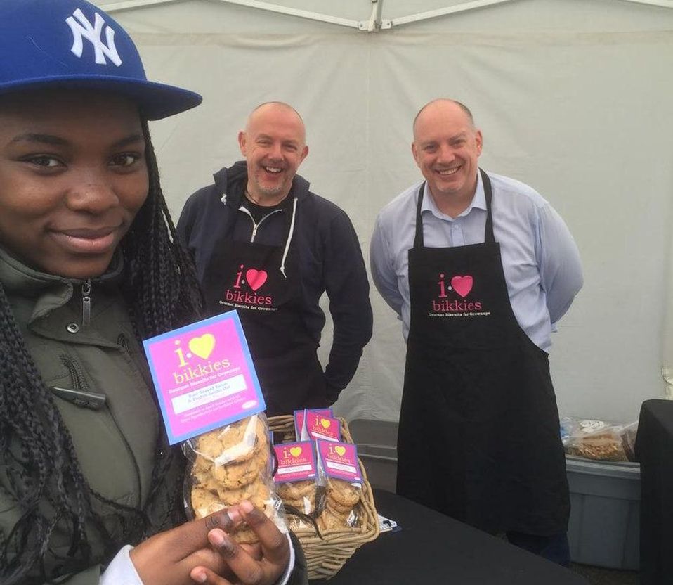Young woman smiles after purchasing gourmet cookies from I Heart Bikkies at a market stall, with two owners